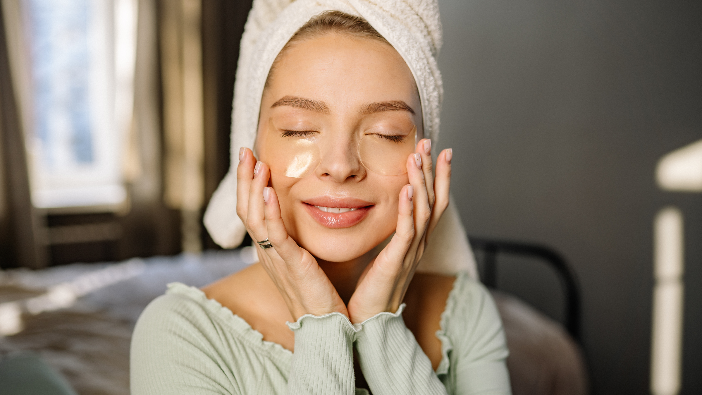 Woman with a towel on her head applying cream to her face in a bedroom setting