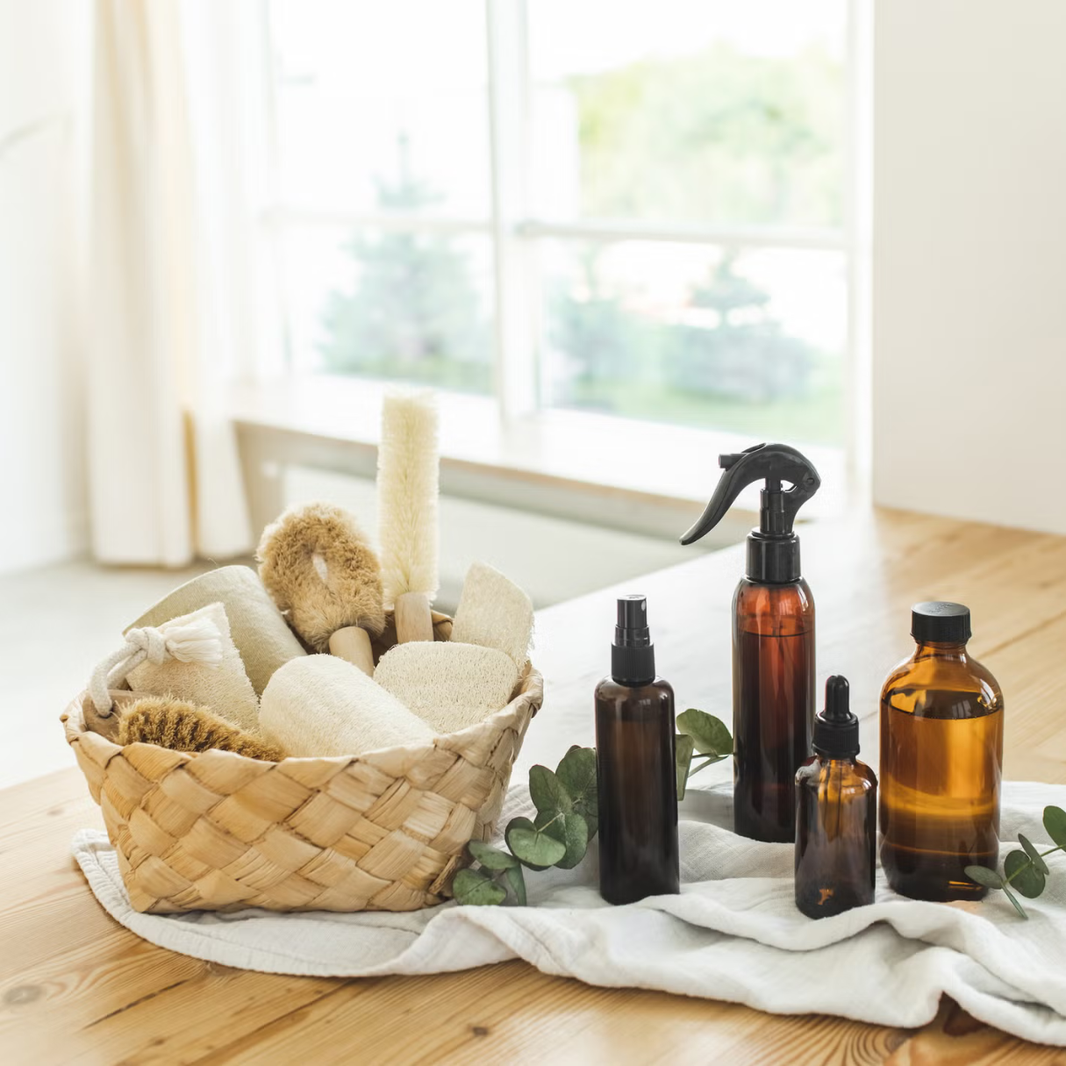 Basket with natural cleaning tools and brown bottles on a wooden surface with a window in the background