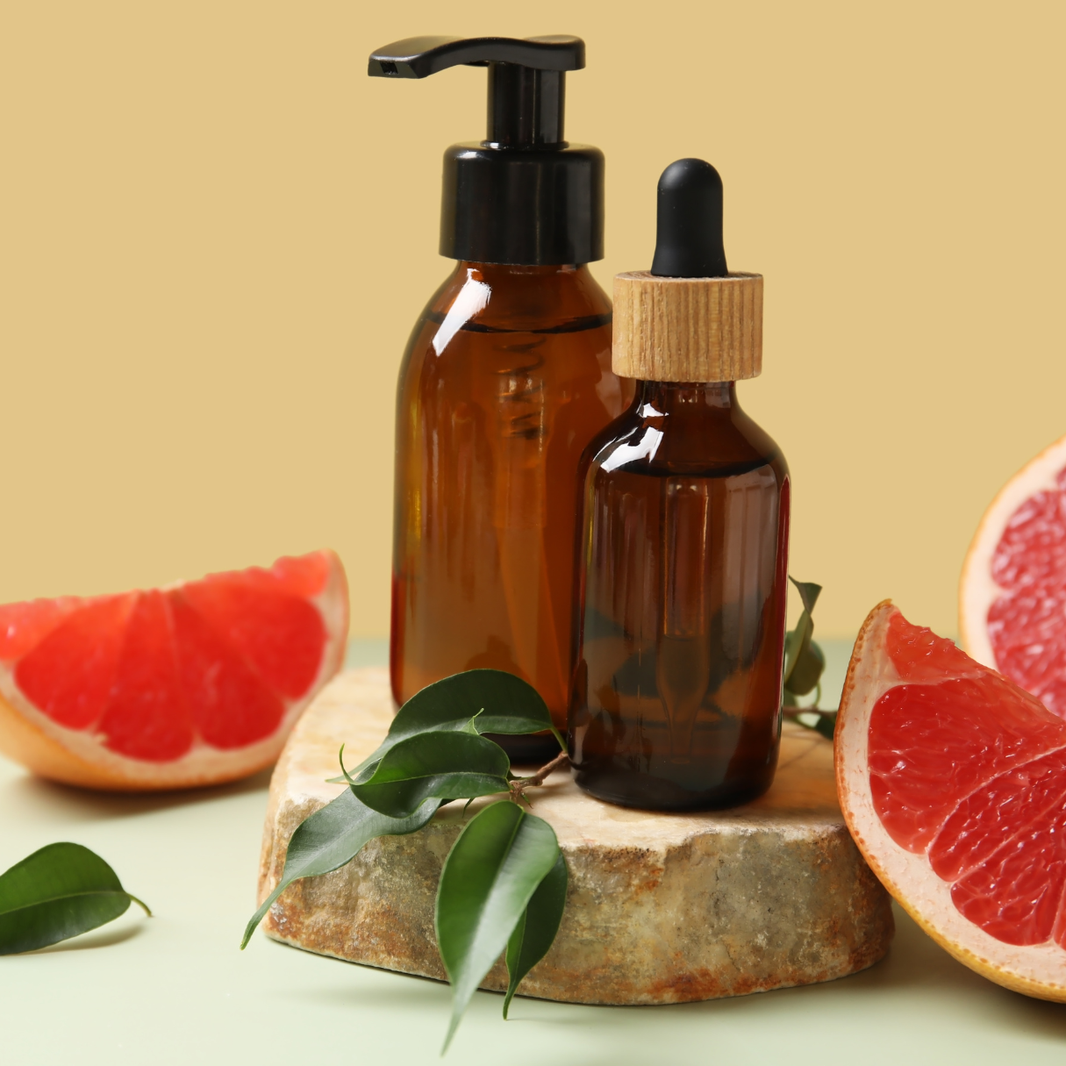 Two brown glass bottles with pump and dropper on a stone surface with grapefruit slices and leaves.