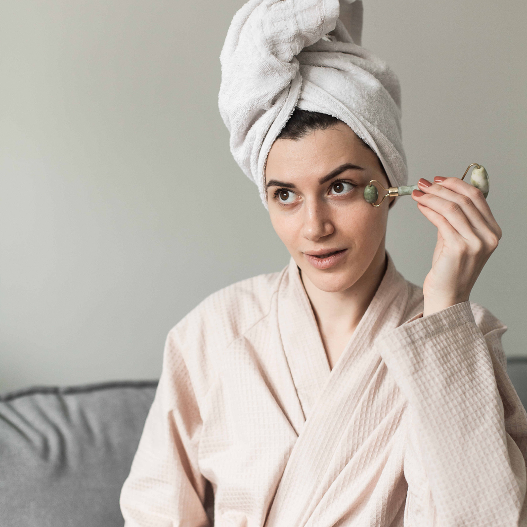 Woman in a robe and towel on head using a facial roller on her face.